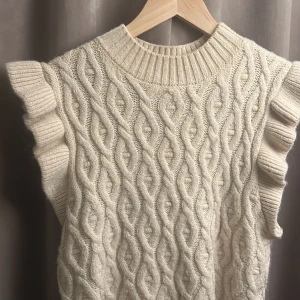 Stickad väst med volang från Name It - Supersöt stickad väst i ljusbeige från Name It. Västen har ett snyggt flätmönster, ribbad hals och nederkant samt breda volangärmar som ger en extra touch. Perfekt att slänga över en skjorta eller t-shirt för en trendig lager-på-lager-look.