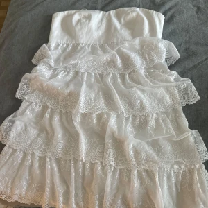 Vit axelbandslös spetsklänning - Supersöt vit axelbandslös kortklänning med flera lager volanger i spets och blommigt mönster. Perfekt för sommarens alla tillfällen när du vill känna dig extra fin och romantisk.