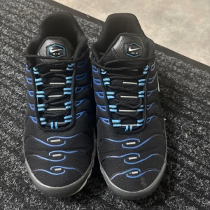 Nike Air Max Plus svarta/blå sneakers - Nike Air Max Plus sneakers i svart mesh med blå och grå detaljer. Skorna har en ikonisk vågig design, vita sulor med synliga Air-enheter och svarta snören med blå inslag. Perfekta för dig som gillar streetwear och vill sticka ut med en klassisk modell. 