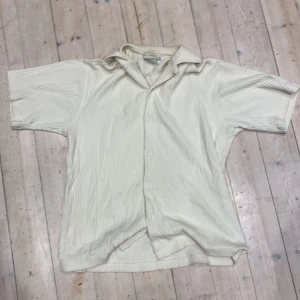Krämvit ribbad skjorta med kort ärm - Säljer en krämvit skjorta med korta ärmar och klassisk krage. Skjortan har en ribbad struktur och knäpps med vita knappar framtill. Materialet känns mjukt och har en lätt textur som ger en cool look. Perfekt för dig som gillar stilrena och enkla plagg.