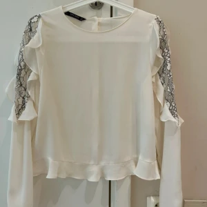 Vit blus med spets och volang Zara - Supersöt  blus från Zara Basic med långa ärmar, volangdetaljer längs axlarna och svart spets på ärmarna. Blusen har knappar i ryggen och en lätt, flowy känsla. Perfekt för dig som gillar romantiska och feminina detaljer.