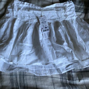 Vit kortkjol med guldprickar - Supersöt vit kortkjol med lågt midjeband och små glittriga guldprickar över hela tyget. Kjolen är lätt och luftig med ett rynkat midjeparti som ger en skön passform. Perfekt för dig som gillar en fräsch och trendig look.