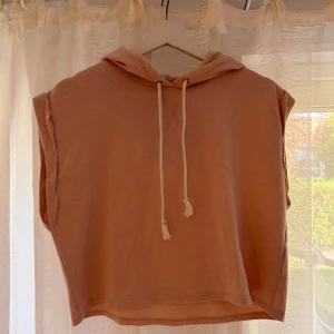 Croppad ärmlös hoodie i ljusrosa - Säljer en croppad hoodie i ljusrosa med huva och snörning framtill. Tröjan är ärmlös och har en avslappnad passform, perfekt för lager-på-lager eller till jeans. Mjuk bomullskänsla och enkel stil som funkar året runt.