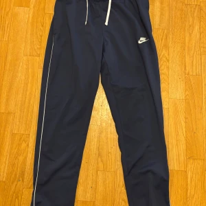 Marinblå Nike mjukisbyxor M - Snygga marinblå mjukisbyxor från Nike med vit logga broderad på låret och vita snören i midjan. Byxorna har en relaxed fit och är gjorda i mjuk polyester. Perfekta för chill eller träning, med diskreta vita detaljer längs benen.