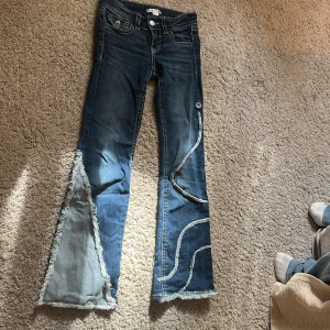 Unika bootcut jeans med fransade detaljer - Säljer ett par blå bootcut jeans med råa, fransade kanter och coola vågformade sömmar på benen. Jeansen har låg midja och extra breda ben med insydda paneler i ljusare denim. Perfekta för dig som vill sticka ut med en edgy och kreativ stil.