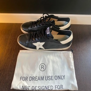 Svarta Golden Goose Superstar sneakers - Golden Goose Superstar sneakers i svart skinn med vit stjärndetalj på sidan och vit häl. Klassisk låg modell med snörning och platt sula. Snygg kontrast mellan svart och vitt, perfekt för dig som gillar streetstyle med premiumkänsla.