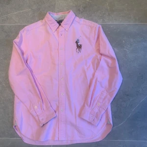 Rosa skjorta från Ralph Lauren storlek L (14/16) passar som S - Snygg rosa skjorta från Ralph Lauren med klassisk button-down krage och broderad polospelare på bröstet. Skjortan har långa ärmar, knappar framtill och är tillverkad i mjuk bomull. Perfekt för dig som gillar stilrena och tidlösa plagg.