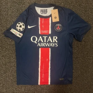 PSG matchtröja D. Doué #14 Nike - Paris Saint-Germain fotbollströja med D. Doué och nummer 14 på ryggen. Mörkblå med röd och vit rand i mitten, officiella klubbmärket och Nike-logga på bröstet. Champions League-märke på ärmen och Qatar Airways som sponsor. Tillverkad i lätt funktionsmaterial.