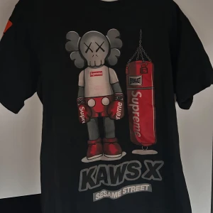 Supreme x KAWS Sesame Street t-shirt - Supreme x KAWS Sesame Street t-shirt i svart med stor grafisk print fram och bak. Framsidan har KAWS-figuren med boxningshandskar och en röd boxningssäck med Supreme-logga. Baksidan har en stor orange KAWS-grafik och figuren. Cool streetwear för dig som gillar unika samarbeten.