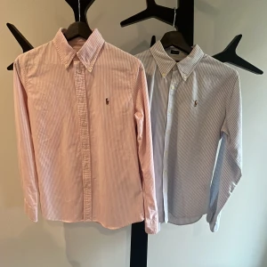 Ralph Lauren Skjortar - Två klassiska skjortor från Ralph Lauren med smala ränder, en i ljusrosa och en i ljusblå. Båda har knappar framtill, lång ärm och broderad logga på bröstet. Perfekt för dig som gillar stilrena och tidlösa plagg. Paketpris 550kr. Styckpris Rosa 400kr Styckpris blå 300kr.