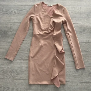 Rosa omlottklänning från Nelly - Säljer en elegant rosa omlottklänning från Nelly. Klänningen har långa ärmar och en v-ringad design med volangdetaljer framtill. Perfekt för en stilren look!