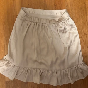 Beige kjol med volang, gjort av silke! - Söt beige kjol med volangdetalj i nederkant. Kjolen har en elastisk midja för bekväm passform och är tillverkad i ett lätt och luftigt material. Perfekt för varmare dagar!
