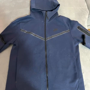 Blå Nike tech fleece - Säljer en snygg blå hoodie från Nike med dragkedja och luva. Den har en diskret logga på bröstet och en praktisk ficka på ärmen. 