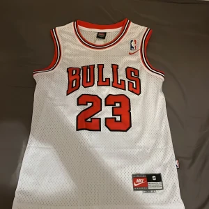Michael Jordan basketlinne från Nike - Säljer ett klassiskt vitt basketlinne från Nike med 'Bulls' och nummer 23 i rött och svart. Linnet har en rund halsringning och ärmlös design, perfekt för basketfans. Detaljerad med 'Jordan' på baksidan. Perfekt för sport eller som samlarobjekt.