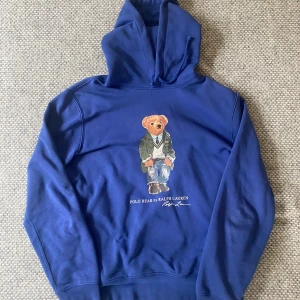 Ralph Lauren Hoodie - Säljer en snygg blå hoodie från Ralph Lauren med en ikonisk Polo Bear-design på framsidan. Perfekt för en avslappnad stil med en touch av lyx. Hoodien har en klassisk passform och är tillverkad i mjukt material för extra komfort. Priset är ej hugget i sten.