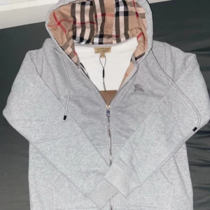 Grå hoodie från Burberry - Säljer en stilren grå hoodie från Burberry med dragkedja och broderad logga på bröstet. Insidan av huvan har det klassiska Burberry-rutiga mönstret. Perfekt för en casual look.