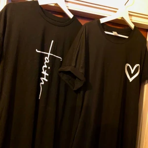 Svart t-shirt med tryck - Två svarta t-shirts med vita tryck. Ena har texten 'faith' i ett korsmönster och den andra har ett hjärta på bröstet. Perfekta för en avslappnad stil.
