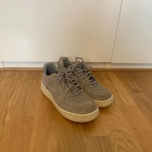 Grå Nike Air Force sneakers - Säljer ett par grå Nike Air Force sneakers i mocka. Skorna har en klassisk design med snörning och rund tå. Perfekta för en stilren och bekväm look.