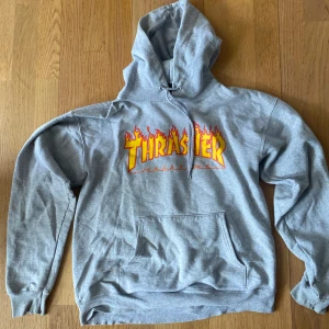 Grå hoodie från Thrasher - Säljer en grå hoodie från Thrasher med den ikoniska loggan i gult och rött med flammor. Tröjan är i bra skick.