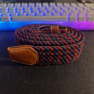 Stilren flätad bälte i marinblått och rostbrunt - Säljer ett knappt använt, elastiskt flätat bälte i marinblått och rostbrunt. Snygg design med läderdetaljer och silverspänne – perfekt för både vardags- och kontorslooken. Bältet är mycket flexibelt och anpassar sig enkelt till din storlek utan förbestämda hål.
