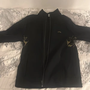 Svart jacka från Boss - Säljer en stilren svart jacka från Boss med dragkedja och diskret broderad logga. Jackan har långa ärmar och en modern design med subtila detaljer i gult. Perfekt för en elegant look. Den är nästan helt oanvänd.