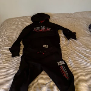 Svart hoodie från Hoodrich - Säljer en svart hoodie från Hoodrich med röd och vit logga på bröstet. Tröjan har en stor ficka framtill och en justerbar huva med dragsko. Perfekt för en avslappnad stil. HOODIEN är M och BYXORNA S.