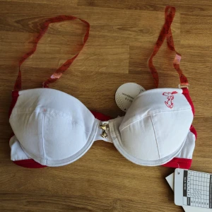 Helt ny vit och röd snobben-bh 65A - Säljer en söt vit och röd bh med ett tryck på Snobben. Bh:n har justerbara axelband i plastliknande material med Snoopy-figurer på. Den är tillverkad i bomull och elastan för en bekväm passform. Den har uttagbara kuddar, är oanvänd och har orginalprislapparna kvar. Perfekt för dig som gillar en lekfull stil! Se också mina andra annonser för möjlighet till att köpa bundles/paket med både rabatt och samfrakt. 