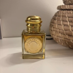 Burberry Goddess Parfym - Elegant parfym från Burberry i en fyrkantig glasflaska med gyllene lock. Flaskan har en stilren design med Burberry-logotypen präglad på framsidan. Perfekt för den som vill ha en lyxig doftupplevelse.