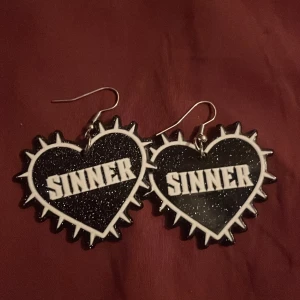 Svarta glittriga örhängen med text - Coola svarta örhängen i form av hjärtan med taggig kant och texten 'SINNER' i vitt. De har en glittrig yta som ger en extra edge till din stil. Perfekta för att göra ett statement!