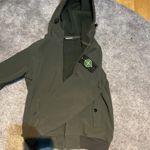 Grön jacka från Stone Island - Snygg grön jacka från Stone Island med huva och dragkedja. Jackan har en ikonisk logga på ärmen och ribbade muddar vid ärmslut och nederkant. Perfekt för en stilren look.