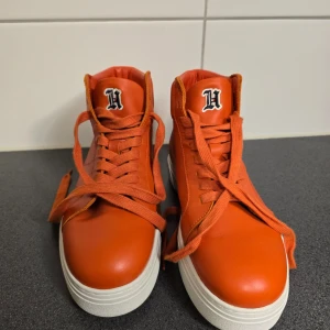 Orange sneakers från Tommy Hilfiger - Snygga orange sneakers från Tommy Hilfiger med hög topp och snörning. Perfekta för en sportig look med en touch av färg. I läder, använda en gång.