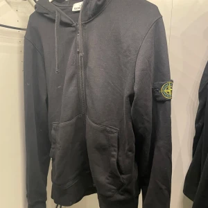 Svart hoodie från Stone Island - Säljer en svart hoodie från Stone Island med dragkedja och det ikoniska märket på ärmen. Tröjan har en justerbar huva med snörning och praktiska fickor framtill. Perfekt för en stilren och avslappnad look. Använd 2 gånger, inga deffekter. Skriv till mig vid frågor eller för mer bilder🤩💫