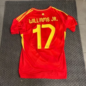 Nico Williams Spanien tröja - Säljer en röd fotbollströja från Adidas med gula detaljer och nummer 17. Tröjan har korta ärmar och är designad för att representera Spanien. Perfekt för fotbollsfans!