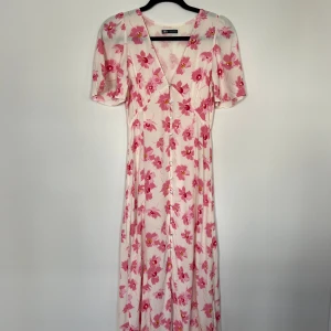 Blommig långklänning från Zara - Säljer en vacker långklänning från Zara med rosa blommönster på en ljus bakgrund. Klänningen har en v-ringad design och korta ärmar, perfekt för sommarens festligheter. Den har knappar framtill som en fin detalj.