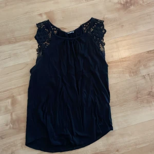 Svart ärmlös topp med spetsdetaljer - Snygg svart ärmlös topp med vackra spetsdetaljer vid axlarna. Perfekt för en stilren look med en touch av elegans. Toppen har en mjuk och följsam passform.