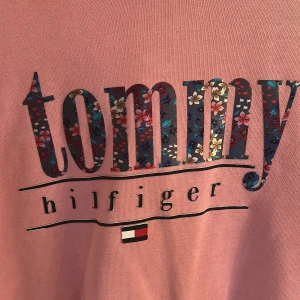Rosa hoodie från Tommy Hilfiger - Säljer en snygg rosa hoodie från Tommy Hilfiger med blommigt mönster i loggan. Tröjan har en klassisk huva och långa ärmar, perfekt för en avslappnad stil. Den har också en praktisk ficka. Perfekt för svala sommar kvällar!
