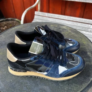 Valentino Rockrunner i blått och kamouflage - Snygga Valentino Rockrunner sneakers i en cool blå och kamouflage design. Skorna har svarta snören och en beige sula som ger en stilren kontrast. Perfekta för dig som vill ha en unik och trendig look.