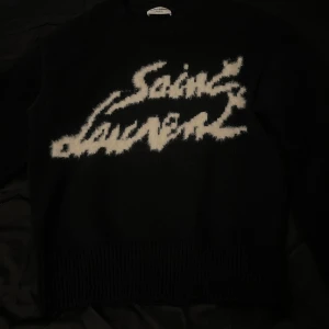 Svart Saint Laurent tröja - Snygg svart tröja från Saint Laurent med vit text på framsidan. Tröjan har en klassisk rund hals och långa ärmar. Perfekt för en stilren look.