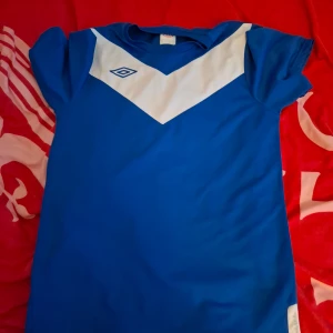 Blå t-shirt från Umbro - Säljer en blå t-shirt från Umbro med vitt V-format mönster på bröstet. Perfekt för sportiga aktiviteter. T-shirten har korta ärmar och är tillverkad i ett lätt och bekvämt material.