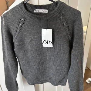 Grå stickad tröja från Zara - Säljer en snygg grå stickad tröja från Zara med coola snörningsdetaljer vid axlarna. Tröjan har en rund halsringning och långa ärmar. Tröjan är helt ny och aldrig använd, prislappar finns kvar. Ursprungs pris var 227 kr.