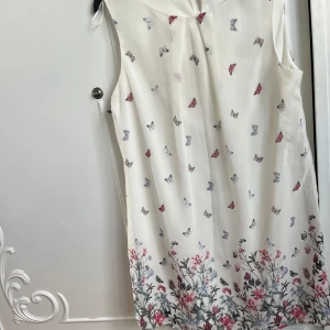 Vit ärmlös klänning med fjärilsmönster - Söt vit klänning med ett färgglatt fjärilsmönster och blommor längst ner. Klänningen är ärmlös och har en luftig passform, perfekt för varma dagar.