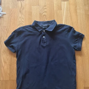 Svart pikétröja från Sail Racing - Snygg svart pikétröja från Sail Racing med klassisk krage och knappar framtill. Perfekt för en stilren look. Märkeslogga diskret broderad på bröstet. Passar både till vardags och mer uppklädda tillfällen.