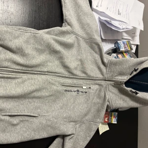 Grå hoodie från Sail Racing helt ny - Snygg grå hoodie från Sail Racing med dragkedja och broderad logga på bröstet. Baksidan har ett stort broderat märke med texten 'Sail Racing Est. 77'. Perfekt för en avslappnad stil. Nypris runt 2000kr