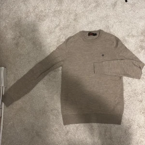 Beige stickad tröja från Morris - Säljer en stilren beige stickad tröja från Morris med ett diskret broderat emblem på bröstet. Tröjan har långa ärmar och ribbade muddar vid ärmslut och nederkant. Perfekt för en avslappnad men ändå elegant look. Nypris på denna är runt 1600kr. Skriv vid funderingar!🙌