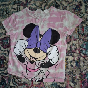 Rosa t-shirt med Musse Pigg från H&M - Säljer en söt rosa t-shirt från H&M med ett stort tryck av Musse Pigg som täcker ögonen med en lila rosett. T-shirten har korta ärmar och ett lekfullt mönster i bakgrunden. Perfekt för barn som älskar Disney!