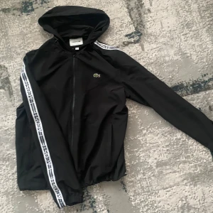 Svart vindjacka från Lacoste - Säljer en stilren svart vindjacka från Lacoste med huva och dragkedja. Jackan har vita detaljer längs ärmarna med Lacoste-logga. Perfekt för en sportig look.