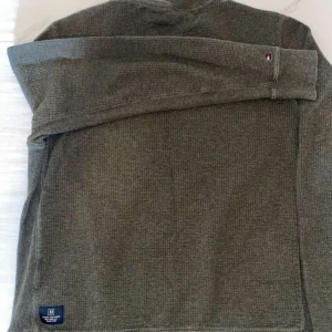 Olivgrön tröja från Tommy Hilfiger - Säljer en olivgrön tröja från Tommy Hilfiger med en unik ficka på bröstet i ett glansigt material. Tröjan har långa ärmar och en rund halsringning. Perfekt för en stilren och avslappnad look.