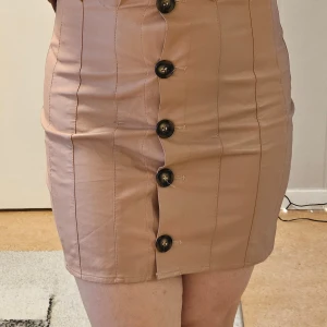 Rosa kortkjol - Snygg rosa kortkjol med framträdande knappar i mitten. Kjolen har en tight passform och är perfekt för en stilren look. Den är gjord i ett material som ger en slät och elegant yta. Knappt använd 