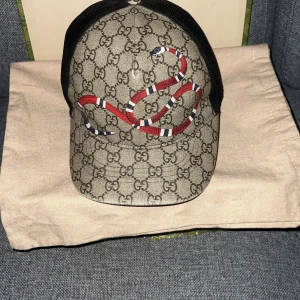 Beige Gucci orm keps - Säljer en snygg äkta beige orm keps från Gucci. Kepsen har en svart baksida och justerbart band för perfekt passform. Perfekt för att ge din outfit en lyxig touch. Kepsen är äkta ,det kan man se på mönstren men kvittot saknas. Påse och box följer med, boxen är lite skadad men det är nästan osynligt.