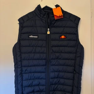 Mörkblå dunväst från Ellesse - Snygg mörkblå dunväst från Ellesse med dragkedja och broderad logga på bröstet. Perfekt för kyligare dagar med sin quiltade design och praktiska fickor. Ett stilrent val för en sportig look. (Helt ny och aldrig använd, lappen sitter fortfarande kvar)